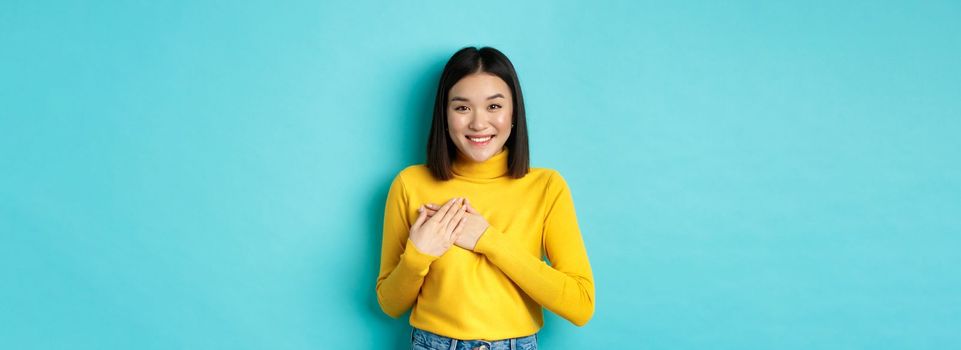 Image of beautiful asian woman holding hands on heart and smiling, thanking you, feeling grateful, standing over blue background.