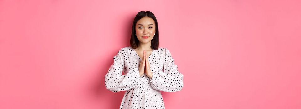Beautiful asian lady in dress asking for help, holding hands in pray or namaste gesture, thanking you, standing over pink background.