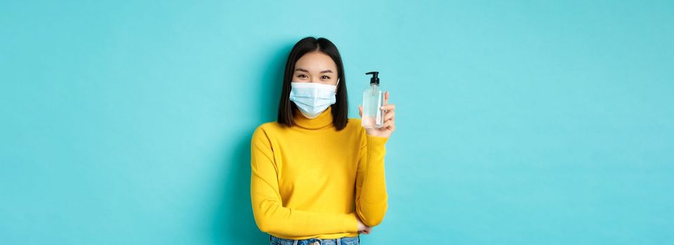 Covid-19, social distancing and pandemic concept. Cheerful asian woman showing hand sanitizer, recommend use antiseptic from coronavirus, wearing medical mask.