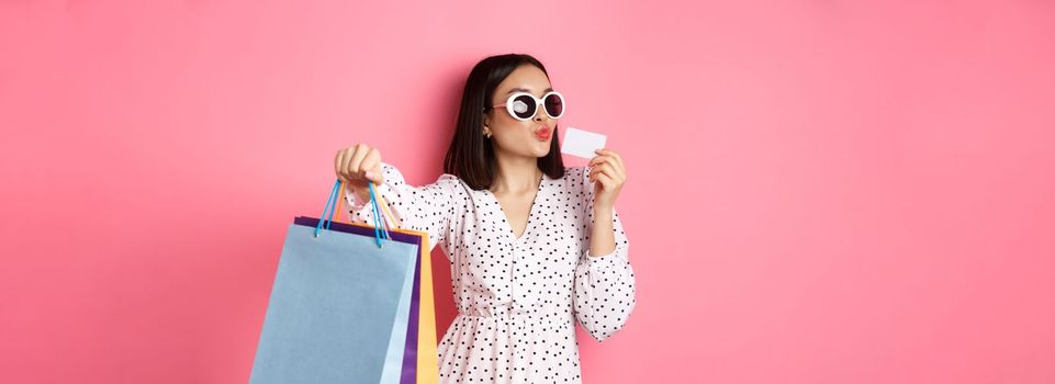 Beautiful asian woman in sunglasses going shopping, holding bags and kissing credit card, standing over pink background.
