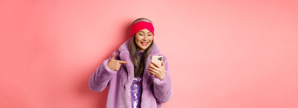 Online shopping and fashion concept. Cool asian senior woman checking out promotion on mobile phone, pointing finger at smartphone screen and smiling pleased, pink background.