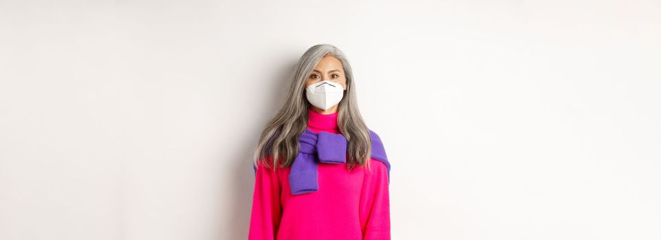 Covid, pandemic and social distancing concept. Stylish asian senior woman wearing respirator and looking at camera serious, standing over white background.