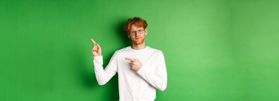 Skeptical caucasian man in glasses having doubts, pouting and frowning while pointing fingers right at something bad, standing over green background.