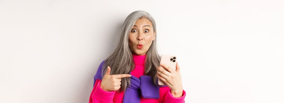 Online shopping. Close up of amazed asian senior woman pointing at smartphone and smiling amazed, standing over white background.