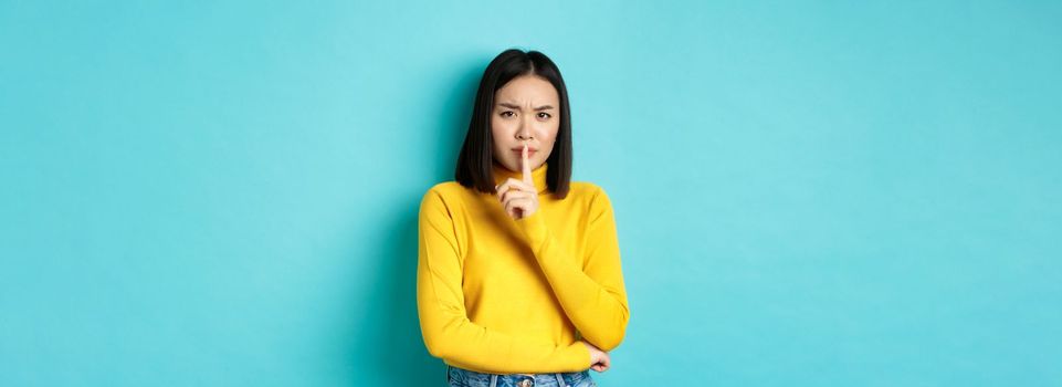 Disappointed asian woman telling to be quiet, scolding loud person with hush gesture, shushing at camera and frowning upset, standing over blue background.