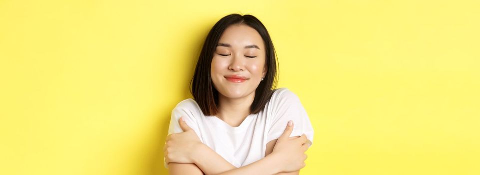 Close up of romantic asian girl hugging herself and dreaming, close eyes and smile while imaging something tender, standing over yellow background.