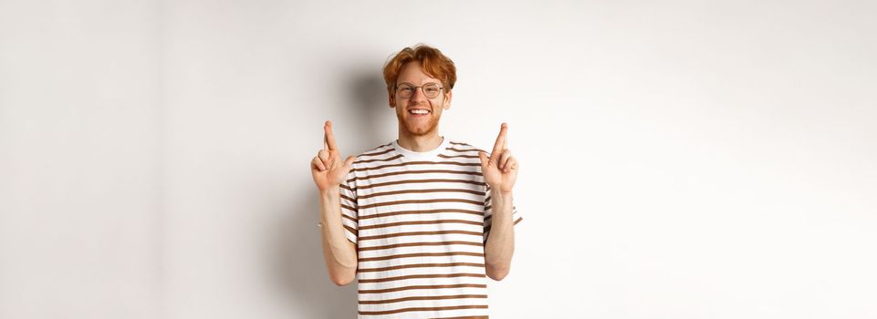 Hopeful and optimistic man with ginger hair and glasses having hope, making wish and cross fingers for good luck, smiling at camera, white background.