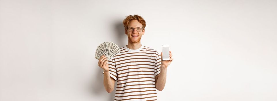 Smiling young redhead man in glasses showing smartphone blank screen and money, standing over white background.