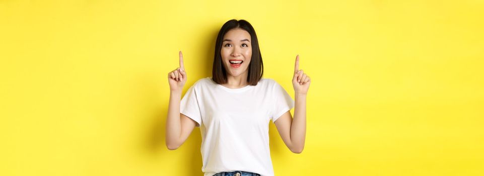 Beauty and fashion concept. Beautiful asian woman in white t-shirt pointing fingers up, standing over yellow background.