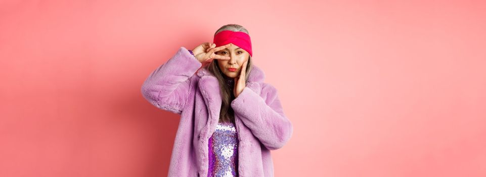 Fashion and shopping. Beautiful asian senior woman in stylish faux fur coat and headband, making peace sign on face and looking sassy and confident at camera, pink background.