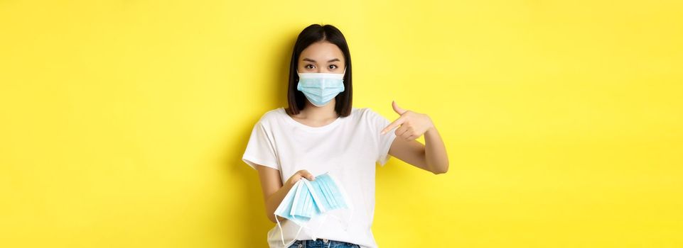 Coronavirus, quarantine and medicine concept. Young asian woman pointing finger at medical masks for going outdoors, standing over yellow background.