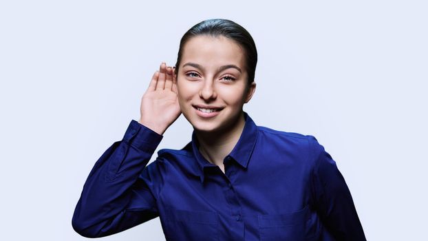 Young attractive teen female holding hand near ear, listening attentively, on white studio background. Secret conversation, news, gossip, eavesdropping concept