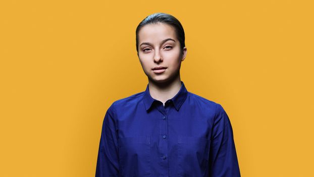 Portrait of serious teenage girl 15, 16 years old, calm young female looking at camera on yellow color studio background. Adolescence, high school education, lifestyle, beauty health concept