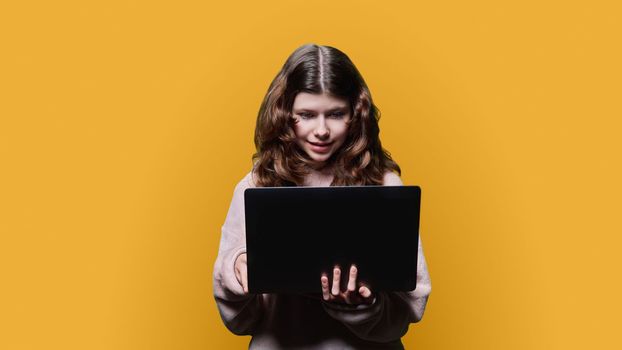 Preteen girl child looking into a laptop, on a yellow studio background. Using computer technology for learning, leisure, childhood adolescence concept