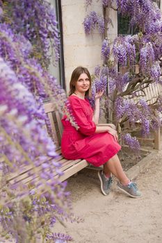 beautiful young woman in red dress sits on a bench in the thickets of blooming wisteria. High quality photo