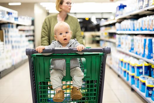 Mother pushing shopping cart with her infant baby boy child down department aisle in supermarket grocery store. Shopping with kids concept