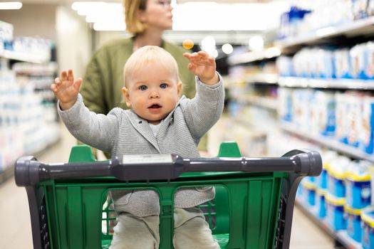 Mother pushing shopping cart with her infant baby boy child down department aisle in supermarket grocery store. Shopping with kids concept