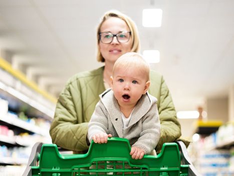 Mother pushing shopping cart with her infant baby boy child down department aisle in supermarket grocery store. Shopping with kids concept