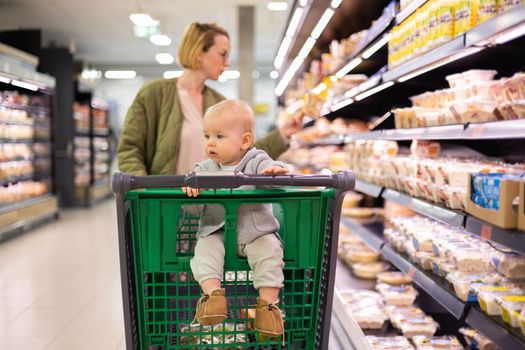 Mother pushing shopping cart with her infant baby boy child down department aisle in supermarket grocery store. Shopping with kids concept