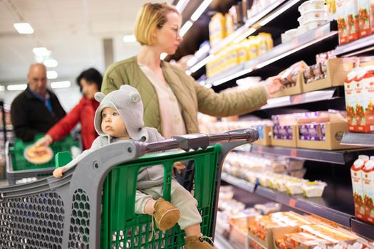 Casualy dressed mother choosing products in department of supermarket grocery store with her infant baby boy child in shopping cart