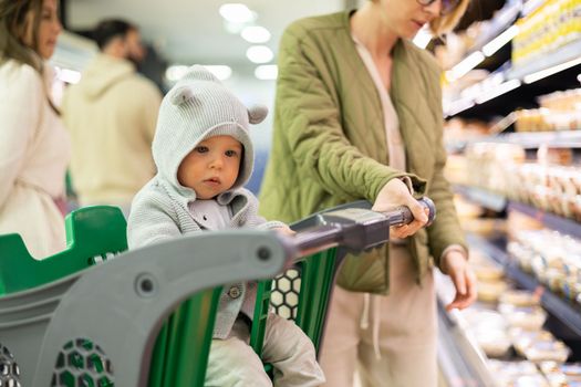 Casualy dressed mother choosing products in department of supermarket grocery store with her infant baby boy child in shopping cart