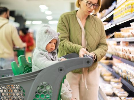 Casualy dressed mother choosing products in department of supermarket grocery store with her infant baby boy child in shopping cart