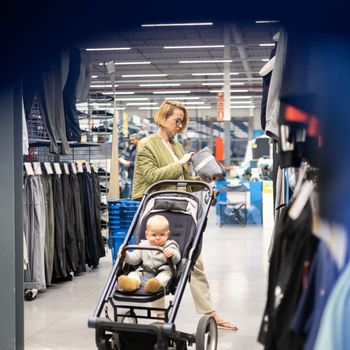 Casualy dressed mother choosing sporty shoes and clothes products in sports department of supermarket store with her infant baby boy child in stroller