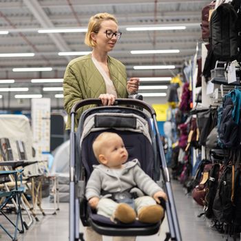 Casualy dressed mother choosing sporty shoes and clothes products in sports department of supermarket store with her infant baby boy child in stroller