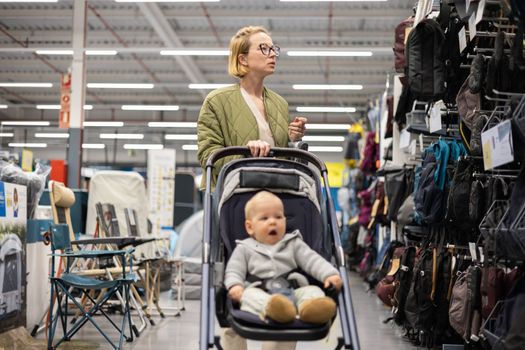 Casualy dressed mother choosing sporty shoes and clothes products in sports department of supermarket store with her infant baby boy child in stroller