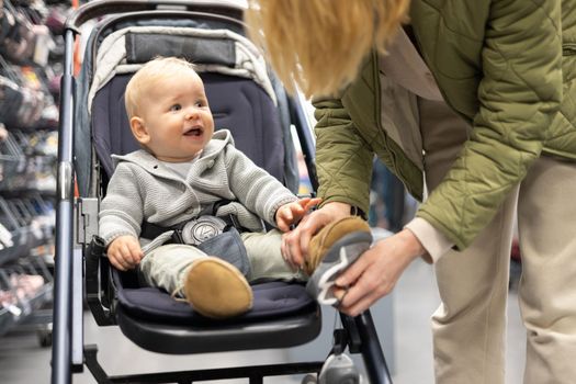 Casualy dressed mother choosing sporty shoes and clothes products in sports department of supermarket store with her infant baby boy child in stroller