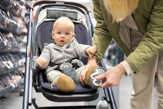 Casualy dressed mother choosing sporty shoes and clothes products in sports department of supermarket store with her infant baby boy child in stroller