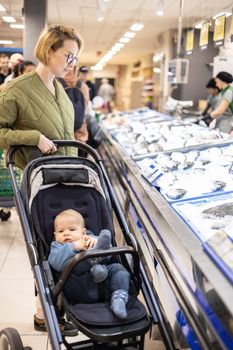 Casualy dressed mother choosing fish in the fish market department of supermarket grocery store with her infant baby boy child in stroller
