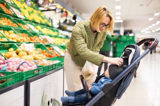 Casualy dressed mother buying fruits and vegetables in supermarket grocery store with her infant baby boy child in stroller