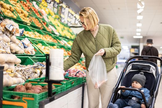 Casualy dressed mother buying fruits and vegetables in supermarket grocery store with her infant baby boy child in stroller