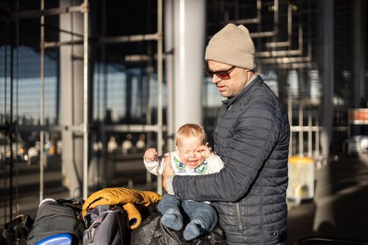 Fatherat comforting his crying infant baby boy child tired sitting on top of luggage cart in front of airport terminal station while traveling wih family