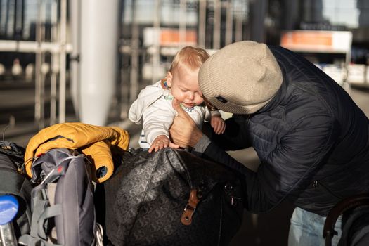 Fatherat comforting his crying infant baby boy child tired sitting on top of luggage cart in front of airport terminal station while traveling wih family