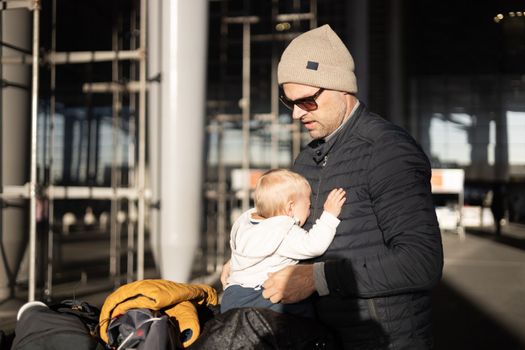 Fatherat comforting his crying infant baby boy child tired sitting on top of luggage cart in front of airport terminal station while traveling wih family