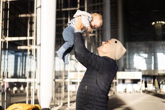 Father happily holding and lifting his infant baby boy child in the air after being rejunited in front of airport terminal station. Baby travel concept