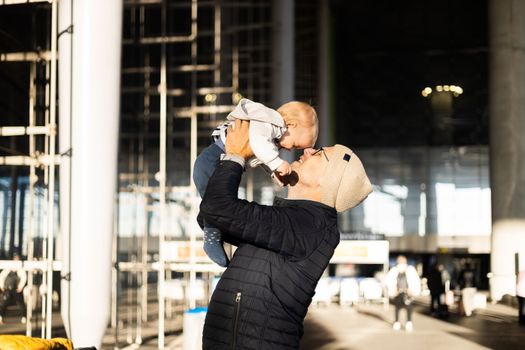Father happily holding and lifting his infant baby boy child in the air after being rejunited in front of airport terminal station. Baby travel concept