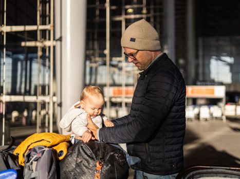 Fatherat comforting his tired infant baby boy child sitting on top of luggage cart in front of airport terminal station while traveling wih family