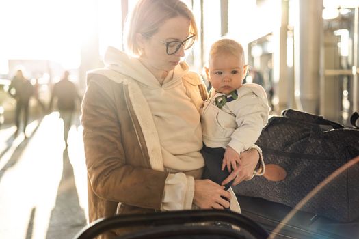 Motherat travelling with his infant baby boy child, walking, pushing baby stroller and luggage cart in front of airport terminal station