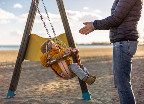 Mother pushing her infant baby boy child on a swing on sandy beach playground outdoors on nice sunny cold winter day in Malaga, Spain