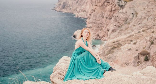 Side view a Young beautiful sensual woman in a mint long dress posing on a volcanic rock high above the sea during sunset. Girl on the nature on overcast sky background. Fashion photo