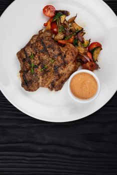 Fried pork chop with grilled vegetables and sauce on white plate on black wooden background