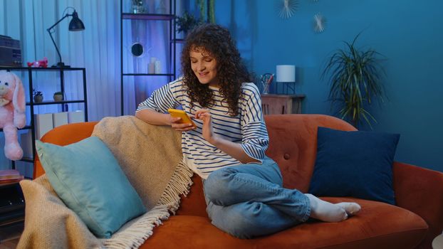Woman with curly hairstyle sitting on sofa uses mobile phone smiles at night home apartment. Girl texting share messages content on smartphone social media applications online, watching relax movie