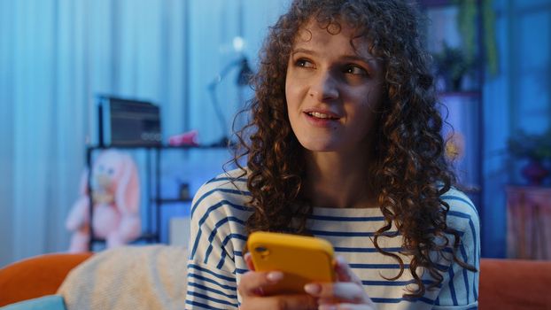 Woman with curly hairstyle sitting on sofa uses mobile phone smiles at night home apartment. Girl texting share messages content on smartphone social media applications online, watching relax movie