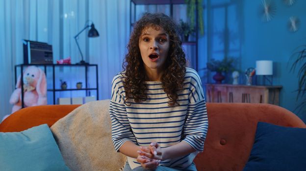 Oh my God, Wow. Surprised woman with curly lush hairstyle at home couch, shocked by sudden victory triumph, good win news, celebrating lottery jackpot luck. Excited amazed girl in night evening room