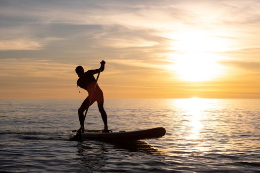 a sporty guy swims with a paddle on a sapboard in the sea under the beautiful sunset sun