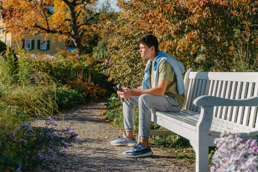 a teenager sits on a bench in the park drinks coffee from a thermo mug and looks into a phone. Portrait of handsome cheerful guy sitting on bench fresh air using device browsing media smm drinking latte urban outside outdoor.