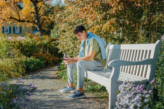 a teenager sits on a bench in the park drinks coffee from a thermo mug and looks into a phone. Portrait of handsome cheerful guy sitting on bench fresh air using device browsing media smm drinking latte urban outside outdoor.
